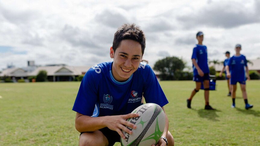 A young boy squats in an oval, holding a rugby ball and smiling at camera