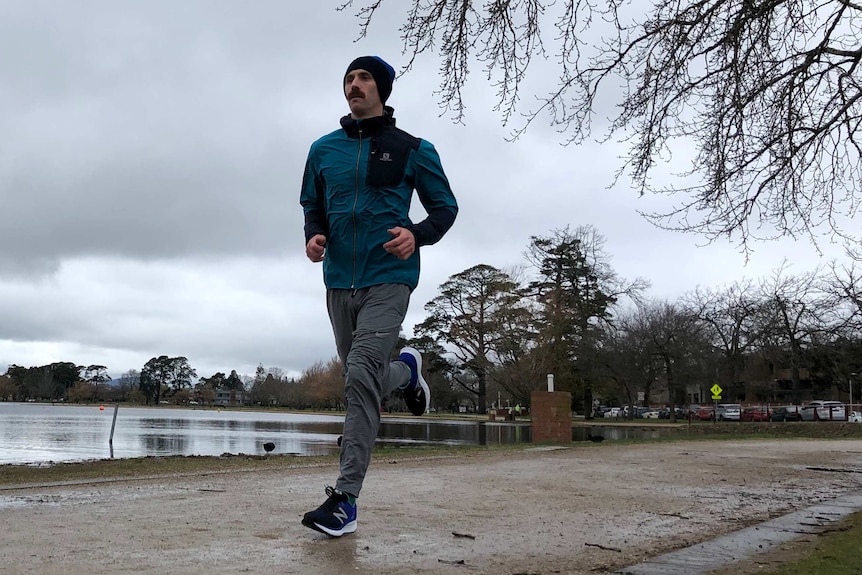 Julian Spence is dressed in a jacket, pants and a beanie as he runs around Lake Wendouree in Ballarat under grey skies.