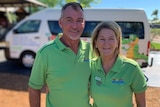 A man and a woman wearing green polo shirts stand smiling posing for a photo in front of a minivan.