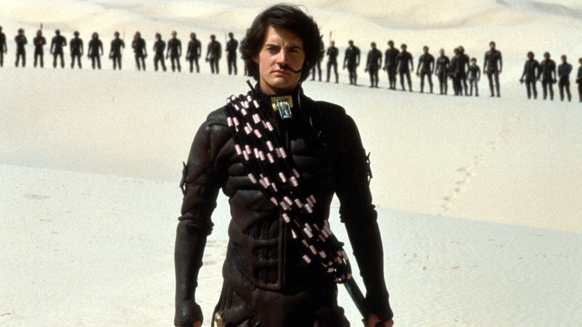 Kyle MacLachlan wearing a black futuristic suit of armour while standing in the desert.