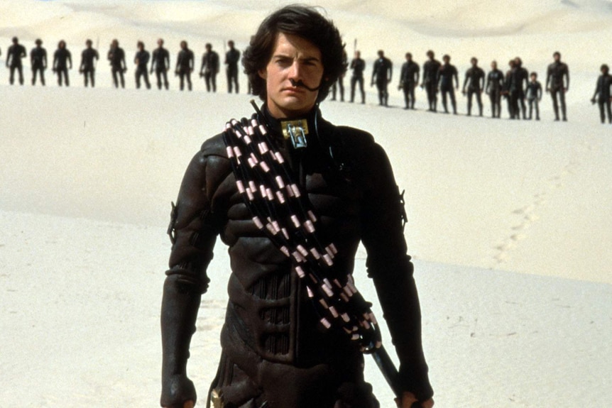 Kyle MacLachlan wearing a black futuristic suit of armour while standing in the desert.