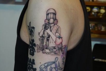 A tattoo on a young man's arm. The image is a man with gas mask holding a camera. An apple is on the side of his body. 