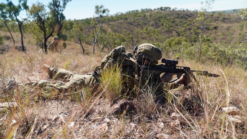 An Australian soldier in battle fatigues lies flat on a grassy knoll aiming his rifle into the distance during training.