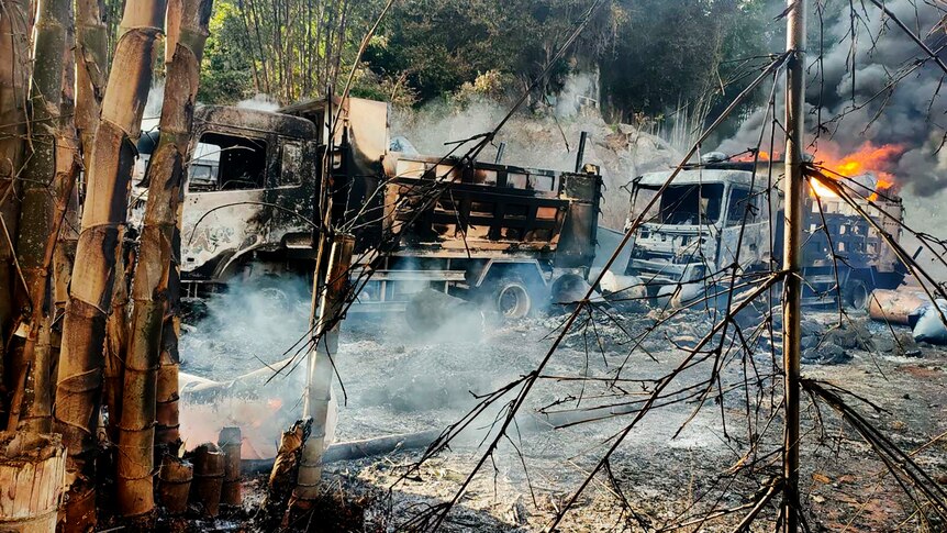 Burnt out vehicles smolder in grassland as flames rise from a truck behind them.