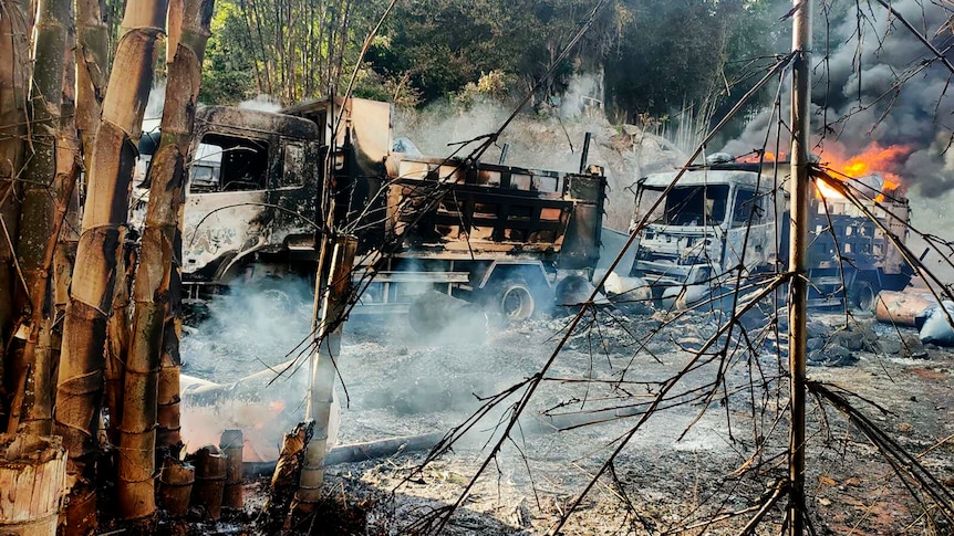 Burnt out vehicles smolder in grassland as flames rise from a truck behind them.
