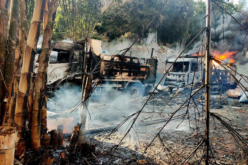Burned-out vehicles smolder in the grasslands as flames rise from a truck behind them.
