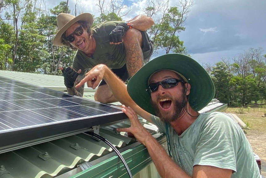 Two men wearing hats smile as they point to a rooftop solar panel.