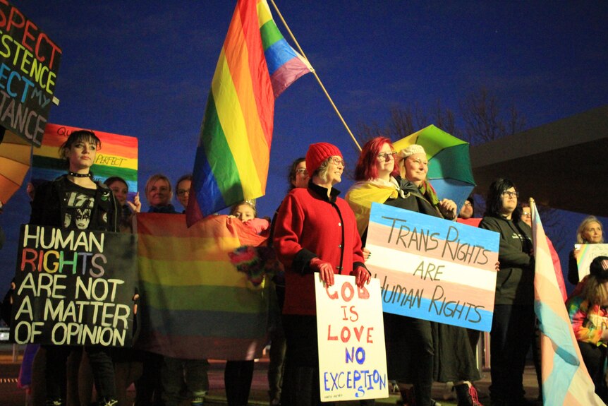 A group of people holding signs and waving LGBTQ flags stand outside a building in the dark. 