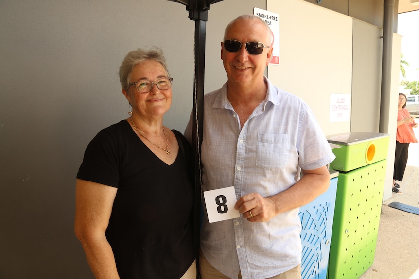 An elderly couple standing in the shade holding the number 8. 