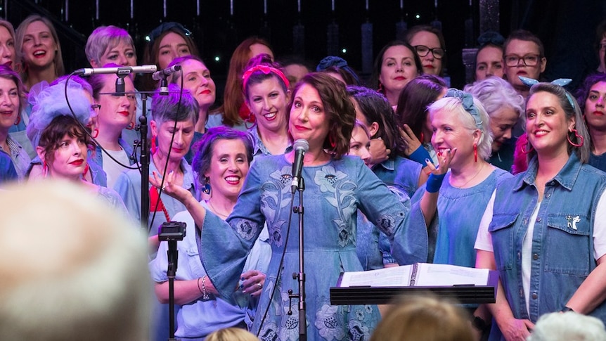 Tamara Oudyn smiles, standing in stage with a choir of women dressed in denim.