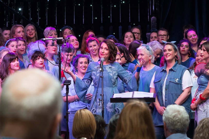 Tamara Oudyn smiles, standing in stage with a choir of women dressed in denim.