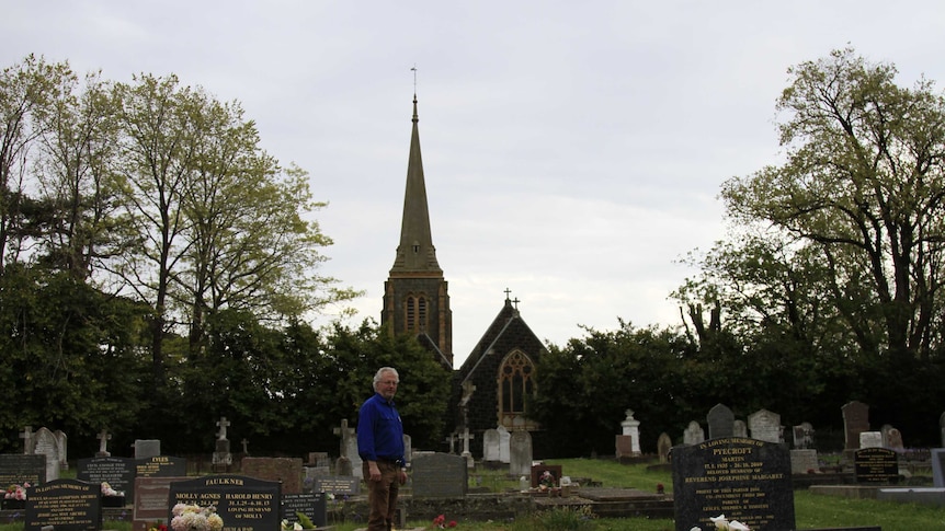 John Temple, a man with white hair and a blue shirt,  in the graveyard of the Anglican Church at Hagley, in Tasmania.