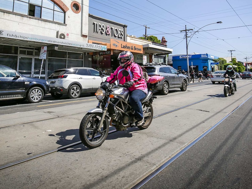 A woman in a pink leather jacket rides down an urban street