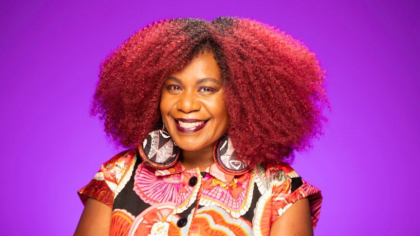 A Black Melanesian PNG woman with a large dark afro with red highlights smiles brightly in front of a purple background