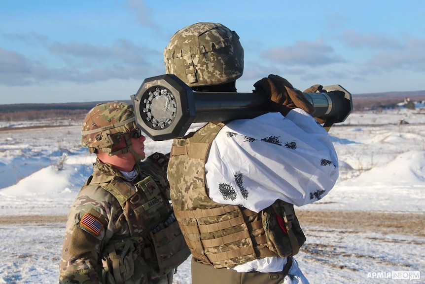 A US instructor in military gear stands behind a Ukrainian soldier holding a M141 Bunker Defeat Munition missile.
