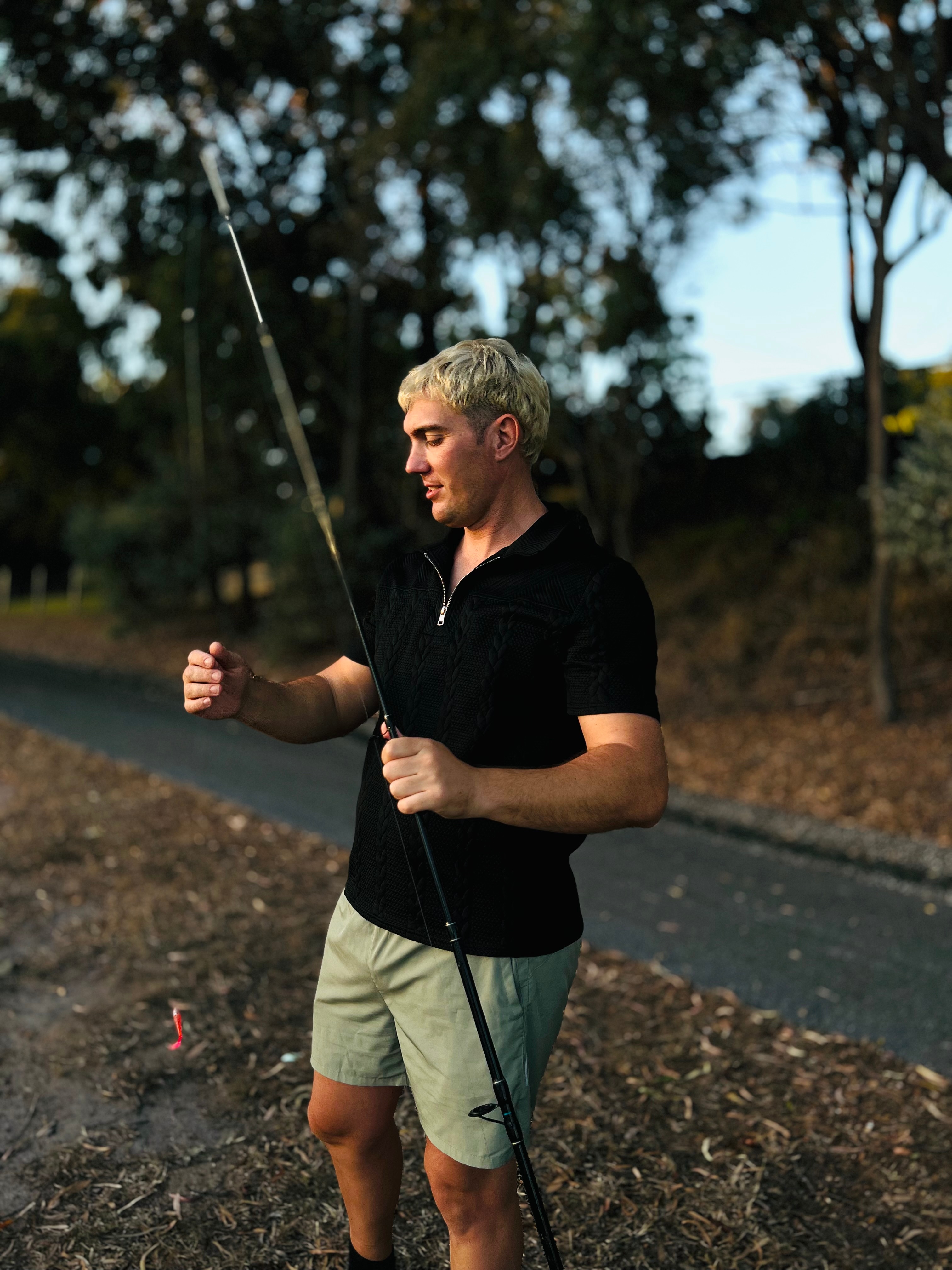 A man stands outdoors by a path. He is looking down at the fishing rod he is holding.