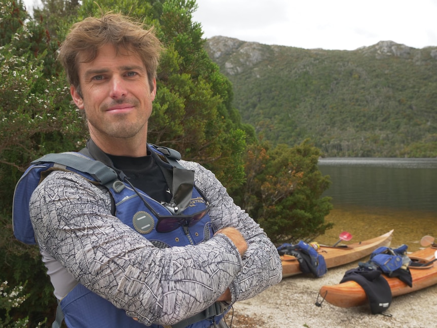 A man in a life vest with crossed arms standing on the shore of a lake, looking to camera, mountains in the background.