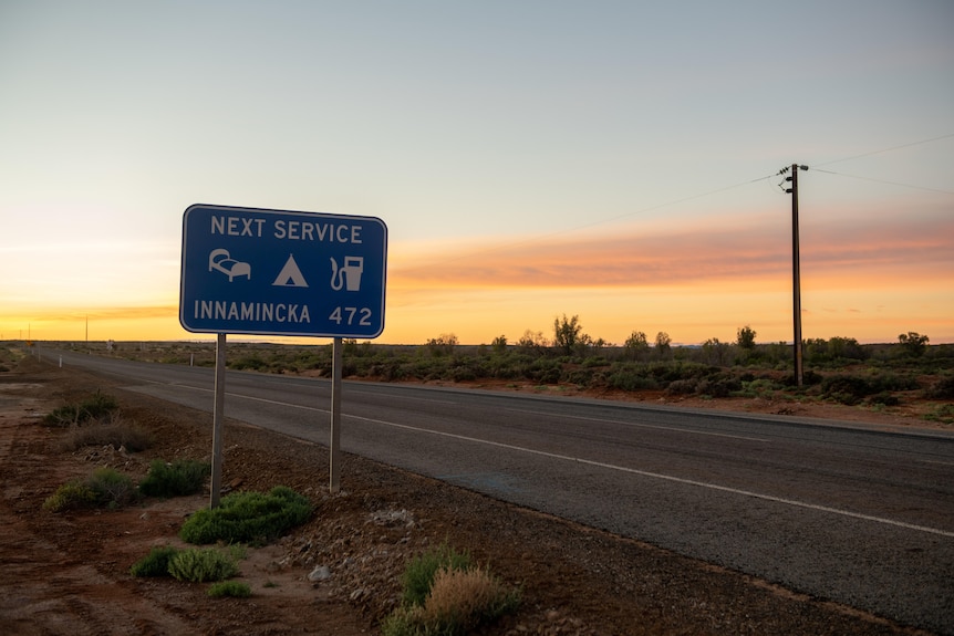 Sunset on a desert road track with road sign that says 472 kilometres to nearest service station. 