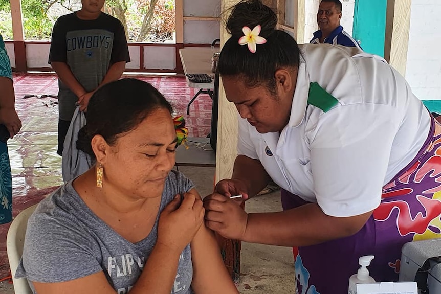 Woman gets vaccinated by another woman with frangipani flower behind her ear.