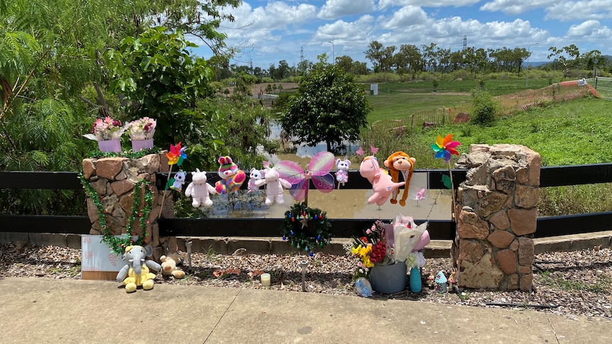 Flowers and stuffed animals marking a memorial at a fence in front of a body of water