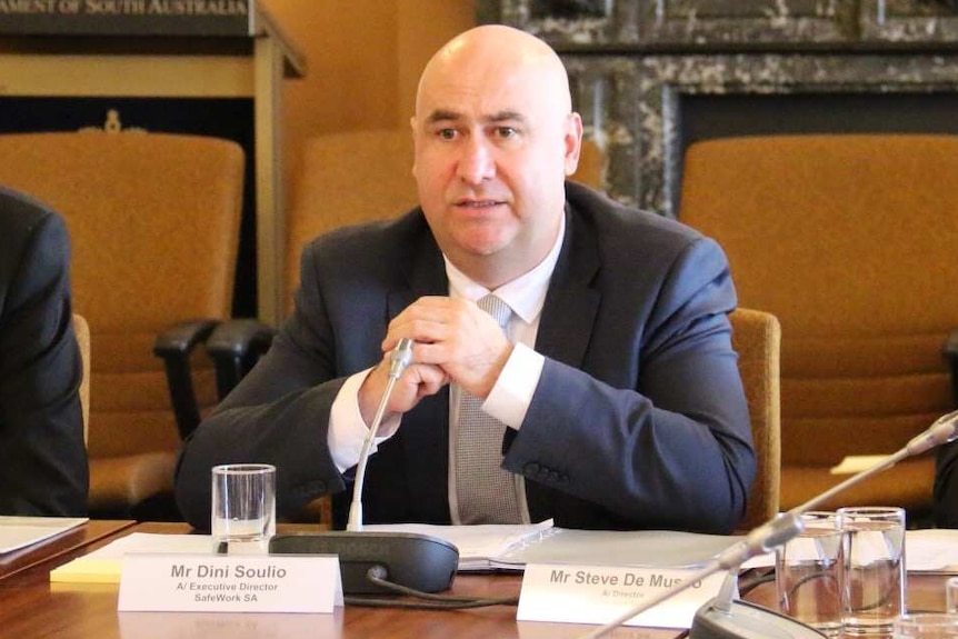 Three men in dark suits sit at a large wooden board table in a conference room.