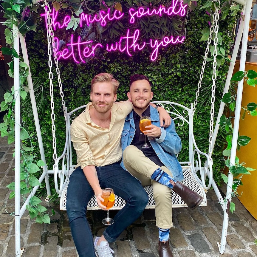 James and Tom pose arm in arm, holding cocktails, under a sign that reads 'the music sounds better with you.'