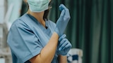 A nurse prepares for surgery wearing blue scrubs and a surgical mask and pulling on a pair of gloves in an operating theatre.