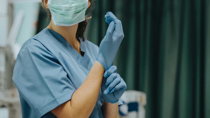 A nurse prepares for surgery wearing blue scrubs and a surgical mask and pulling on a pair of gloves in an operating theatre.