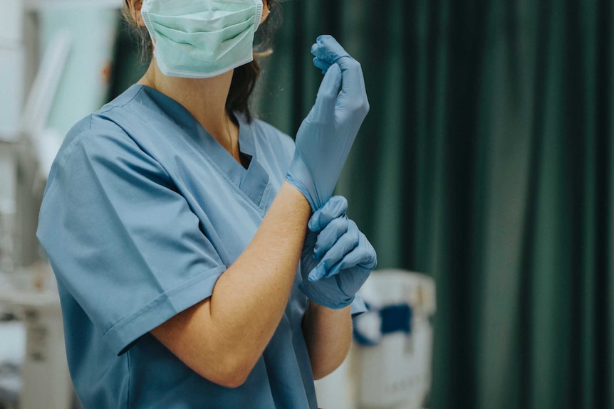 A nurse prepares for surgery wearing blue scrubs and a surgical mask and pulling on a pair of gloves in an operating theatre.