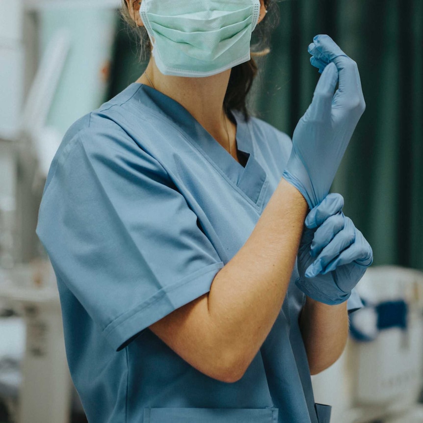 A nurse prepares for surgery wearing blue scrubs and a surgical mask and pulling on a pair of gloves in an operating theatre.