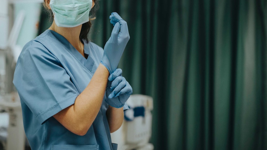 A nurse prepares for surgery wearing blue scrubs and a surgical mask and pulling on a pair of gloves in an operating theatre.