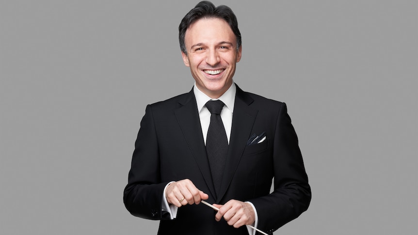 Conductor Umberto Clerici in a black suit, smiling and holding his baton against a grey background