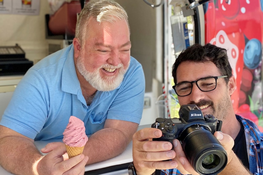 One man stands in an icecream van smiling, looking at the back of an SLR camera that another man is holding, he's also smiling