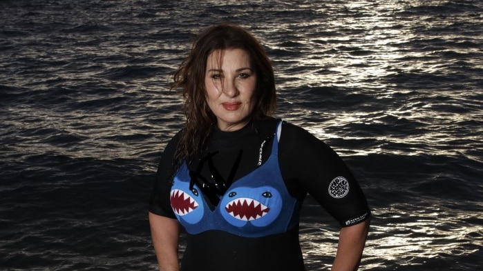 A brown haired woman stands in shallow water, wearing a wetsuit and a bra with sharks painted on them over the top.