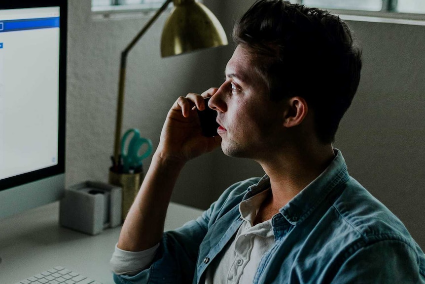 A man talks on the phone while sitting at his home desk staring at his computer monitor