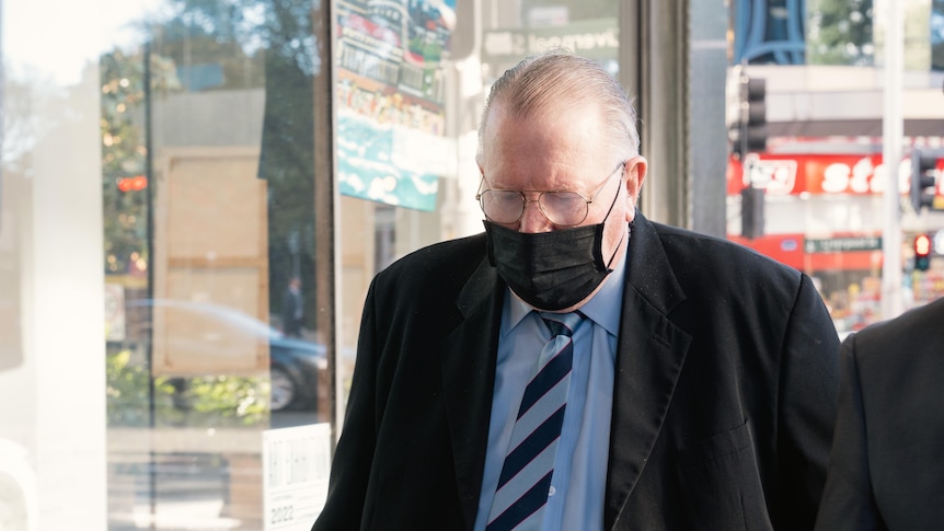 elderly man wearing a black face mask, glassed and suite and tie, walks forward with his head down