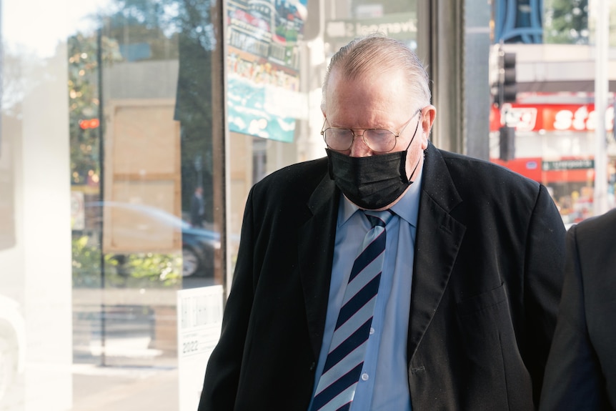 elderly man wearing a black face mask, glassed and suite and tie, walks forward with his head down