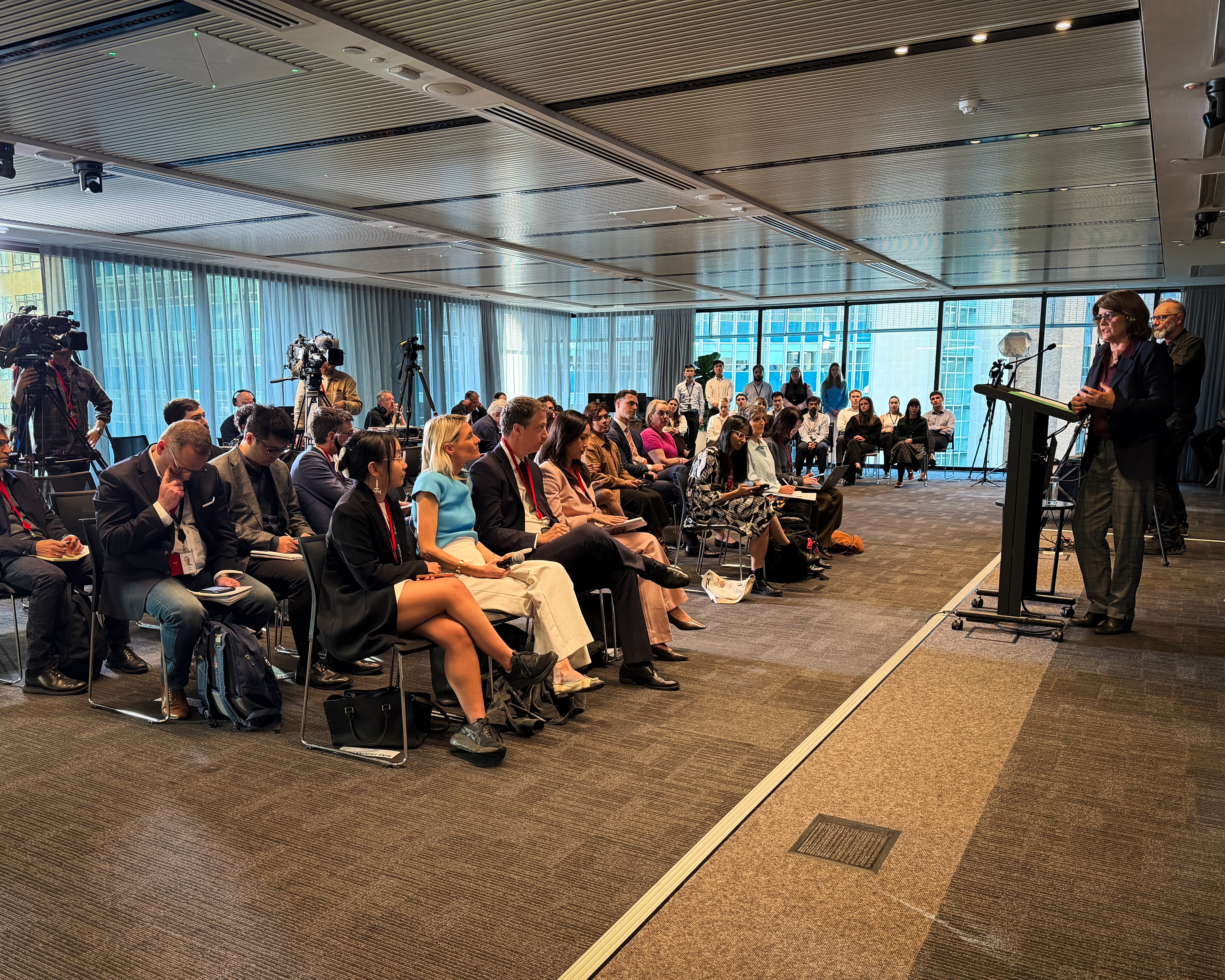 A woman stands at a lectern and speaks to journalists in chairs in a room with floor-to-ceiling windows.