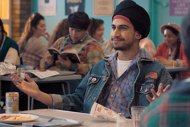 Aaron Phagura wearing blue denim jacket with American flag patch smiles and gestures with arms open at cafeteria lunch table.