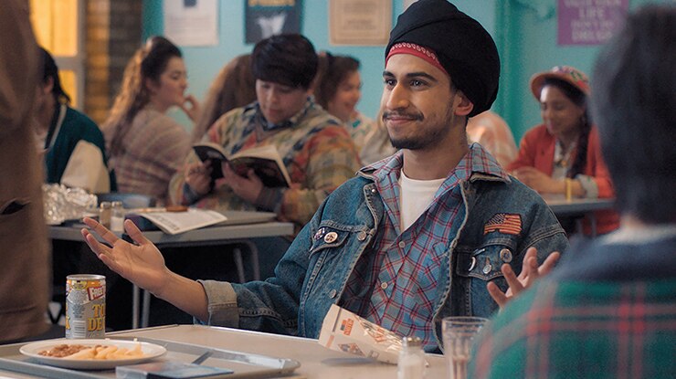 Aaron Phagura wearing blue denim jacket with American flag patch smiles and gestures with arms open at cafeteria lunch table.