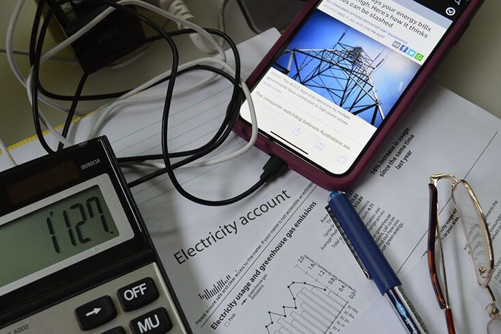 A flat lay of a calculator, an electricity bill and a mobile phone displaying an ABC article about electricity prices. 