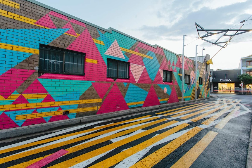 A pink, green and yellow abstract mural, metal sculpture, and yellow and white road markings, in Byron Bay's Lateen Lane.