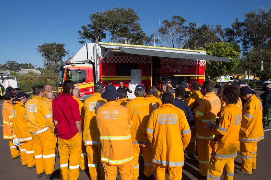 A fresh team of firefighters clump together in their orange uniforms to be briefed in front of fire truck.