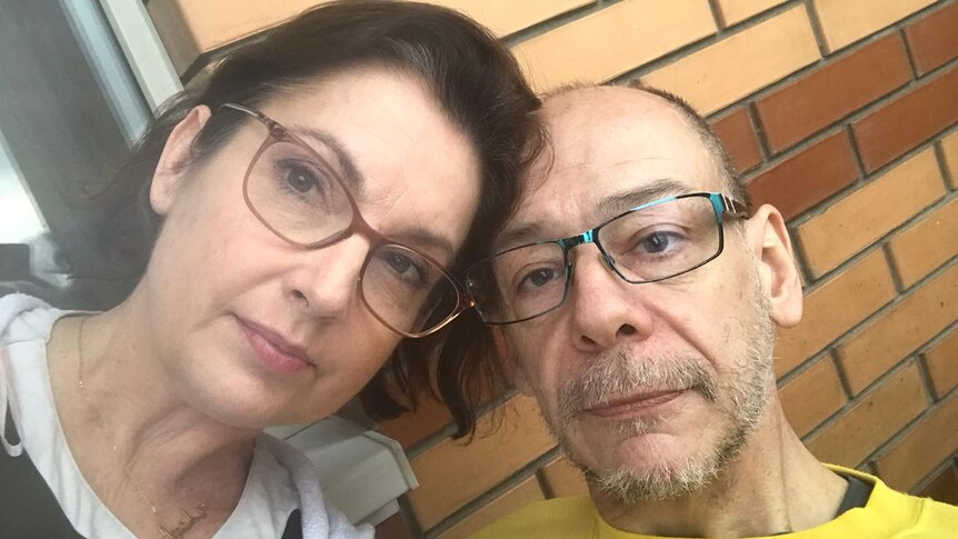 A woman with brown hair and glasses with a bald man with glasses against a brick wall balcony.