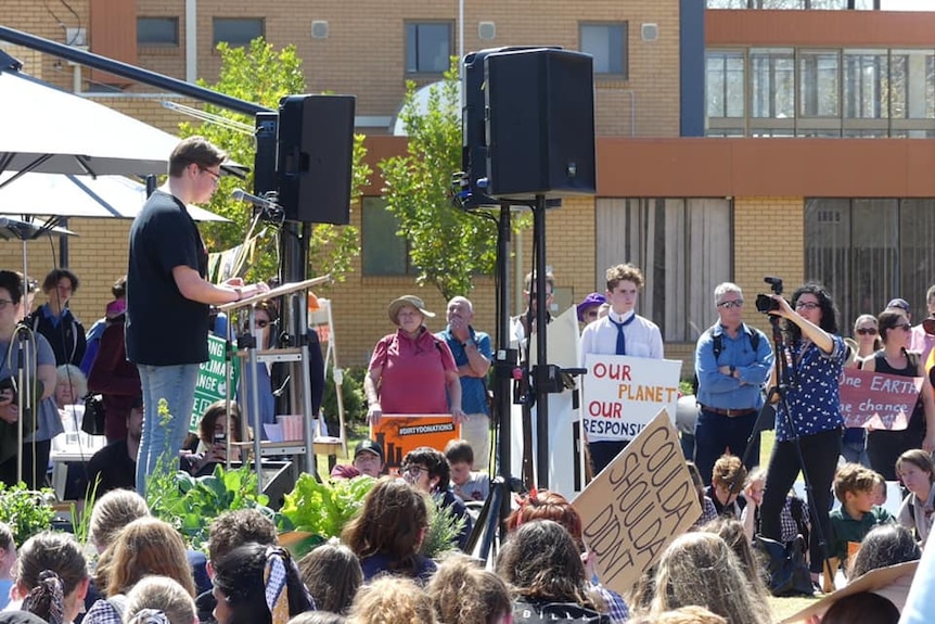 A teenage boy stands on a stage in a park and addresses the crowd