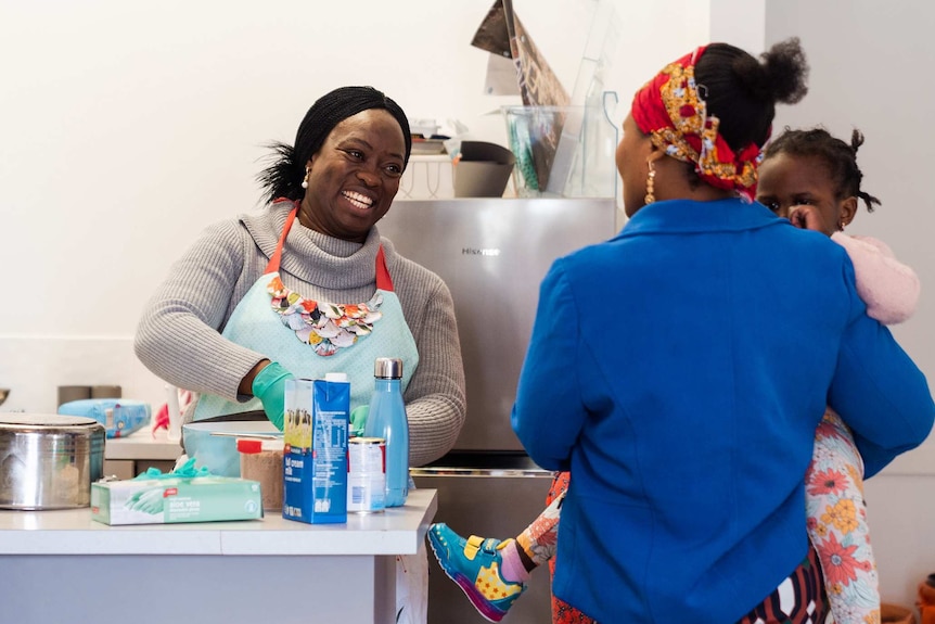 A woman stands at a bench in a kitchen mixing food in a bowl and smiling as she talks to another woman who is holding a baby.