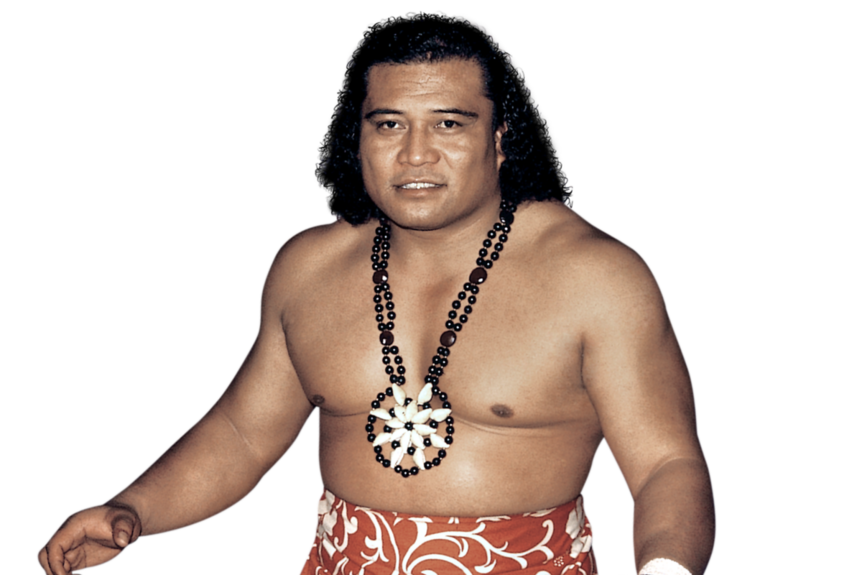 Male wrestler with black shoulder length curly hair stands with red ie lavalava and black and white neck piece. 