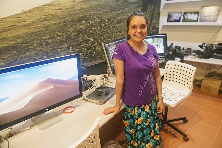 Sienna Stubbs stands in a purple shirt leaning against a desk with three computers on it.