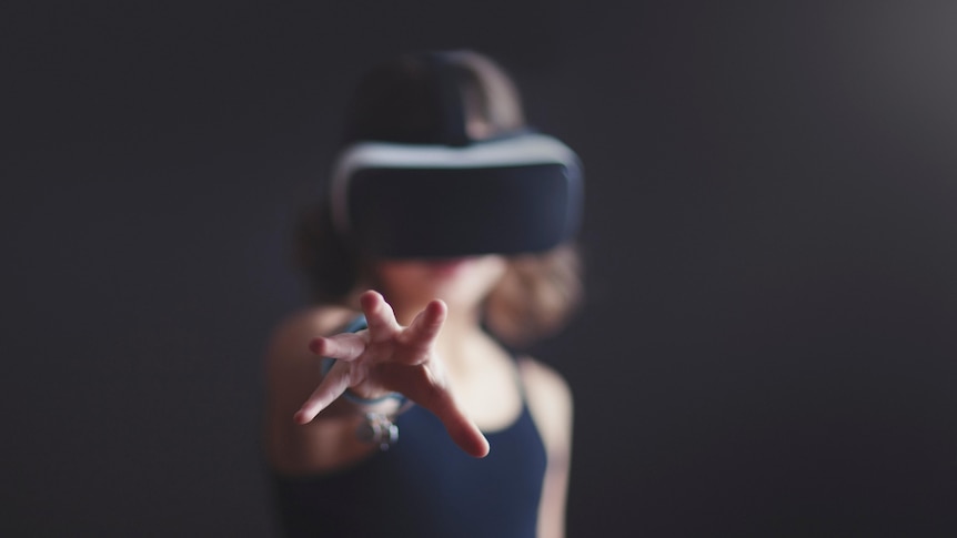 An image of someone wearing a VR headset. They're in front of a black background and are reaching forward towards the camera.