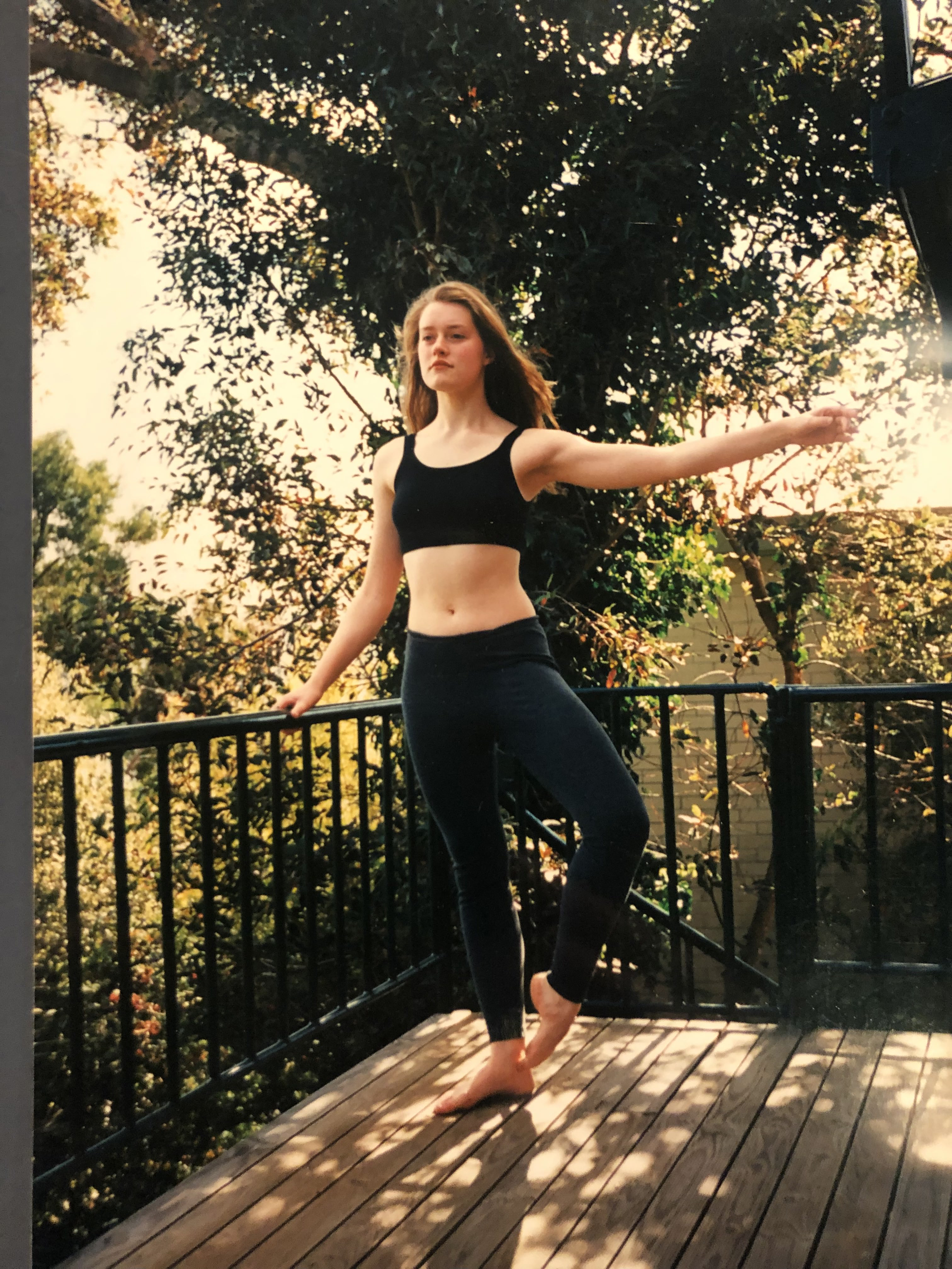 White teenaged girl with mousey brown hair wears black active wear and holds arms in a ballet port de bras on an outdoor deck.